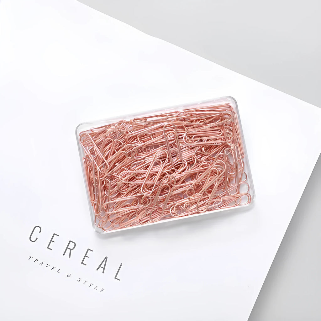 Box of 100 small rose gold heart-shaped metal paper clips on a white surface.