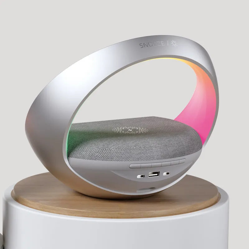 Beige multifunctional wireless charger clock with Bluetooth speaker and LED wake-up light on a bedside table.
