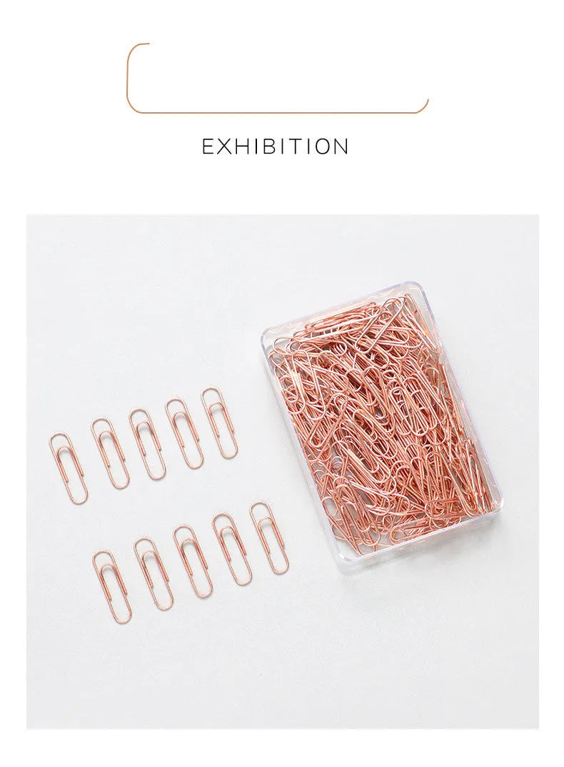 Box of 100 small rose gold heart-shaped metal paper clips on a white surface.
