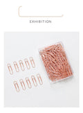 Box of 100 small rose gold heart-shaped metal paper clips on a white surface.