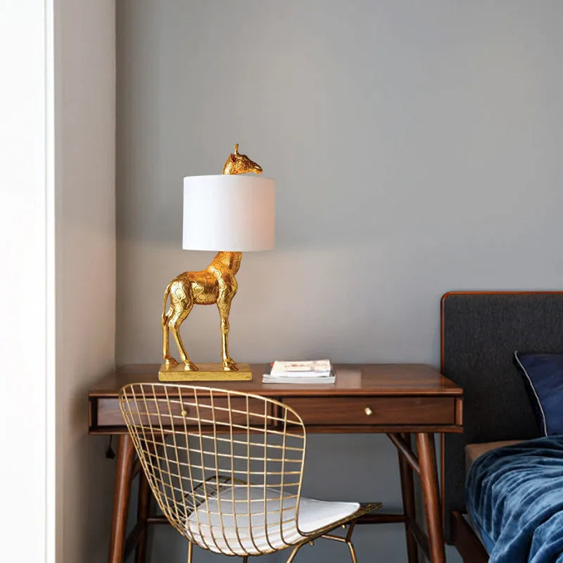 A tall white giraffe table lamp with a cloth shade on a wooden desk.