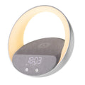 Beige multifunctional wireless charger clock with Bluetooth speaker and LED wake-up light on a bedside table.