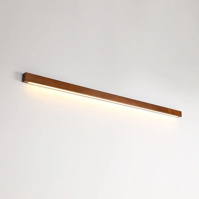 Long, slim walnut-coloured wooden wall lamp with integrated LED lighting on a plain wall.