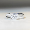Close-up of a silver geometric ring with a twisted band and a 1-karat moissanite stone in a four-claw setting.