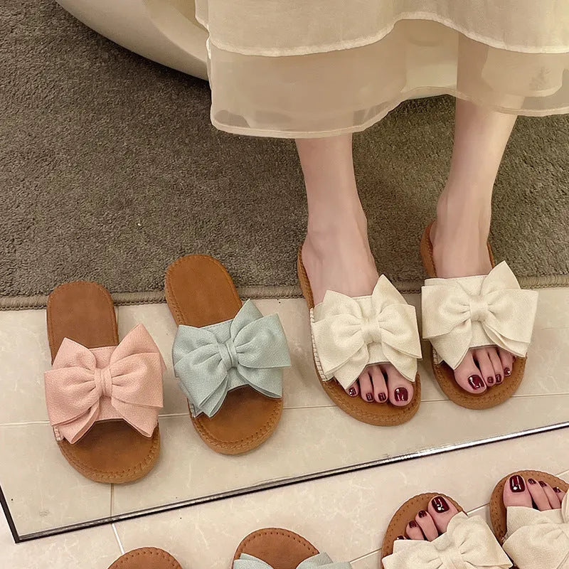 Women's beige slip-on sandals with a bow detail, suede cloth upper, and soft rubber flat sole.