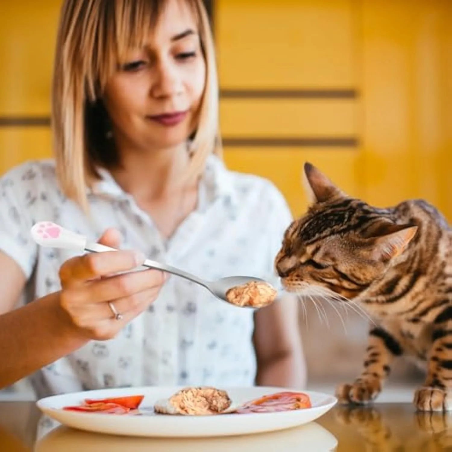 White and black stainless steel pet food spoons with silicone cat paw print handles on a table.