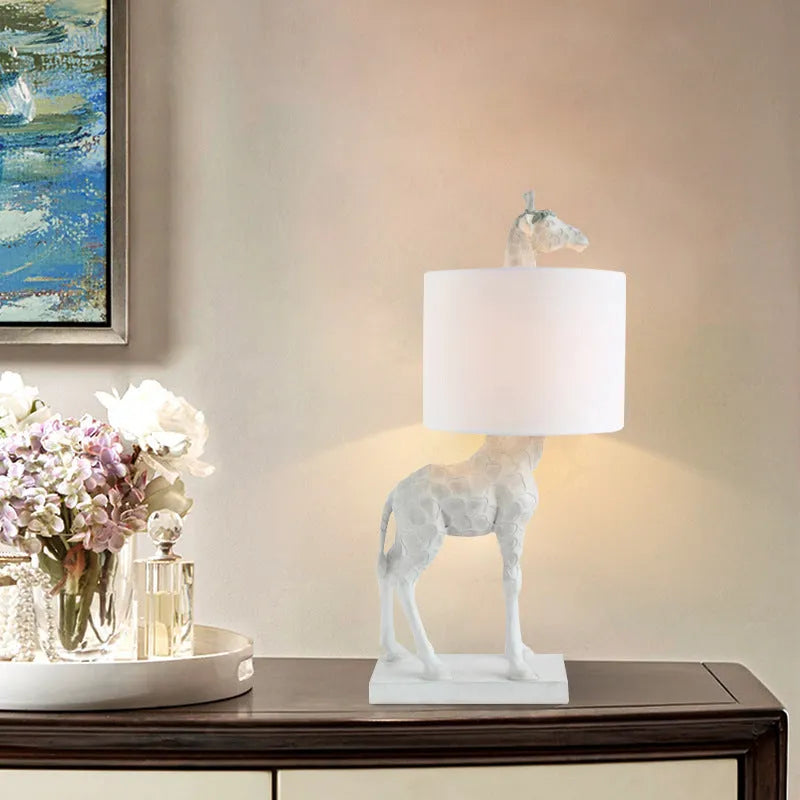 A tall white giraffe table lamp with a cloth shade on a wooden desk.
