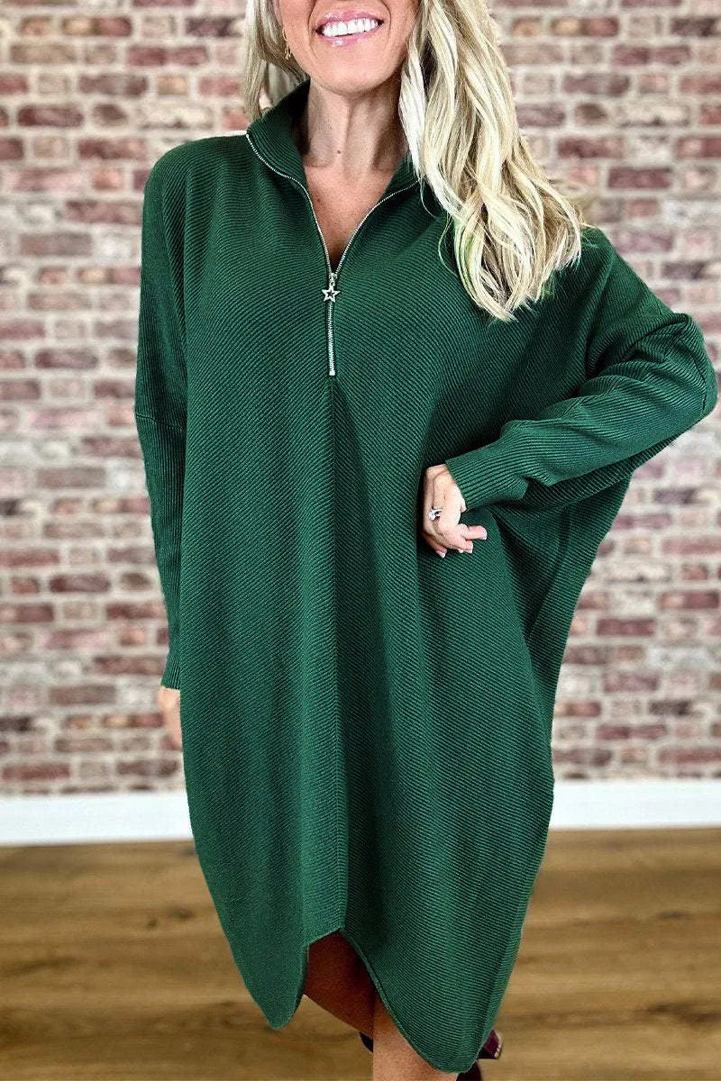 A woman wearing a solid dark green loose-fit knitted dress with raglan sleeves and a zipper detail.