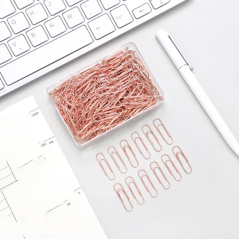 Box of 100 small rose gold heart-shaped metal paper clips on a white surface.