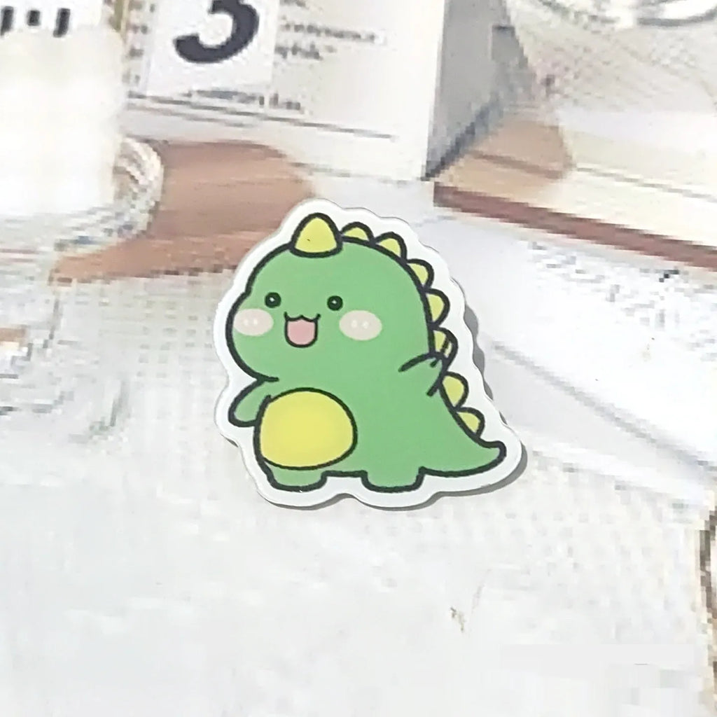 Cartoon dinosaur acrylic badge brooch with printed design and pin fastening, shown in one of twelve colour styles