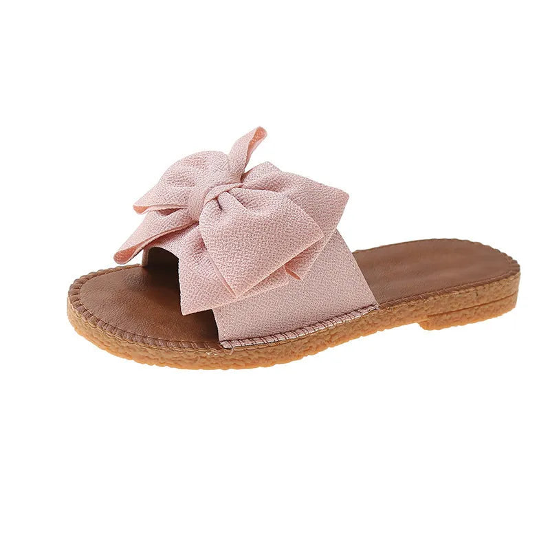Women's beige slip-on sandals with a bow detail, suede cloth upper, and soft rubber flat sole.