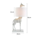 A tall white giraffe table lamp with a cloth shade on a wooden desk.