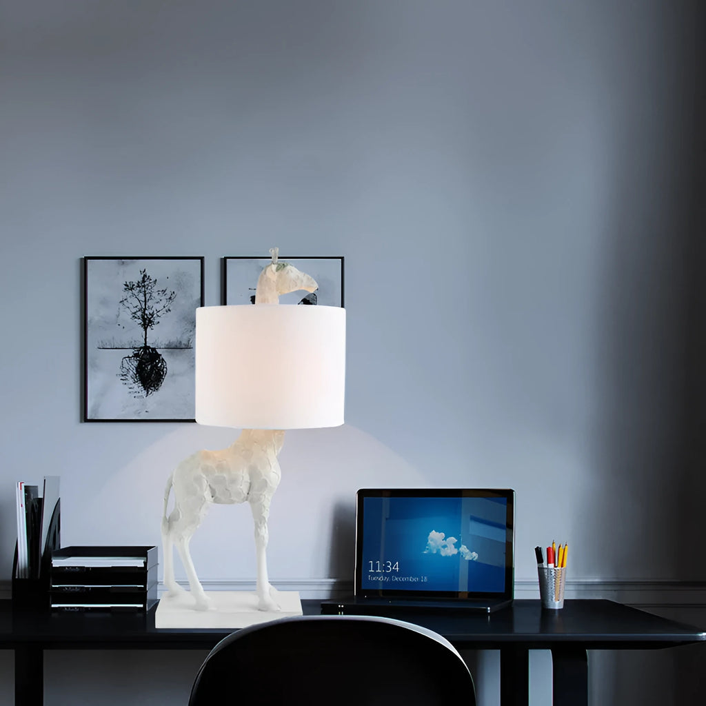 A tall white giraffe table lamp with a cloth shade on a wooden desk.