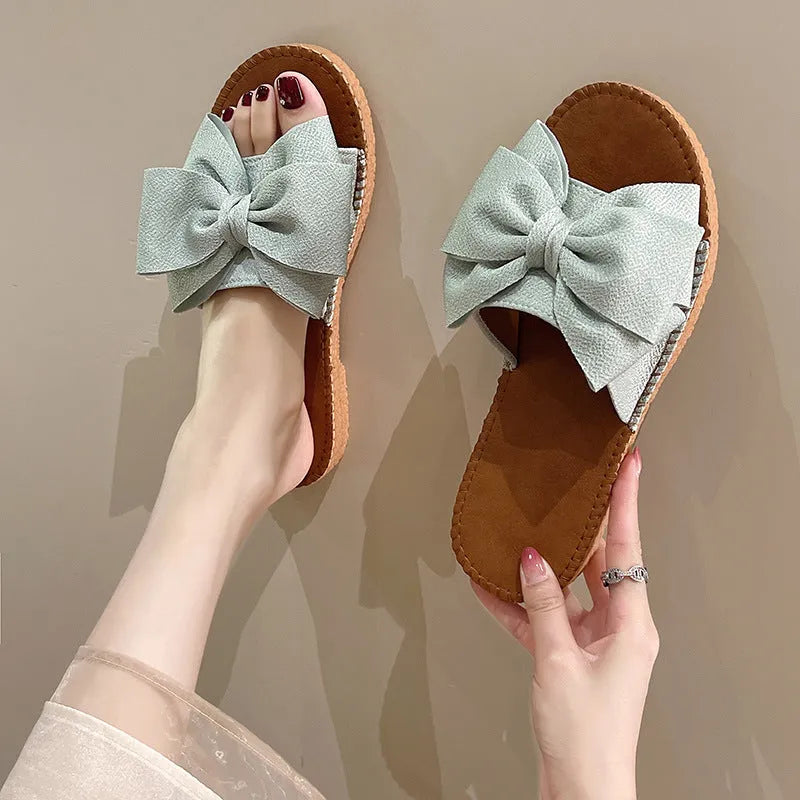 Women's beige slip-on sandals with a bow detail, suede cloth upper, and soft rubber flat sole.