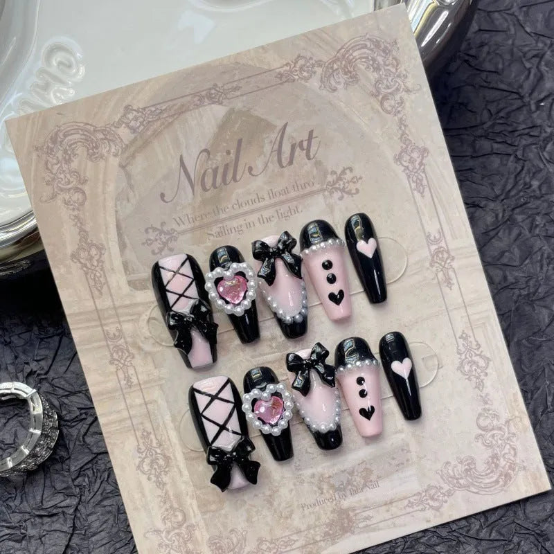 Black and pink heart-shaped hand-painted false nails with bow design, shown in mid-length cat's eye style.